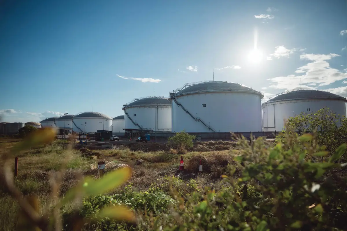 Large white industrial storage tanks at an energy facility, surrounded by vegetation under a bright blue sky.