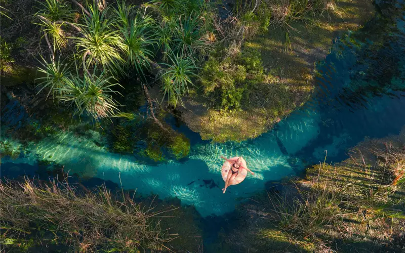 Person laid back and relaxing on an inflatable in the clear blue waters of Bitter Springs surrounded by lush tropical vegetation