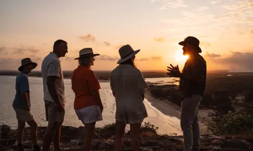 A group of five visitors stands on a coastal lookout at sunset, overlooking a winding tidal waterway and expansive landscape. One person appears to be guiding or speaking to the group while the setting sun casts the figures in silhouette against the warm orange and golden sky.