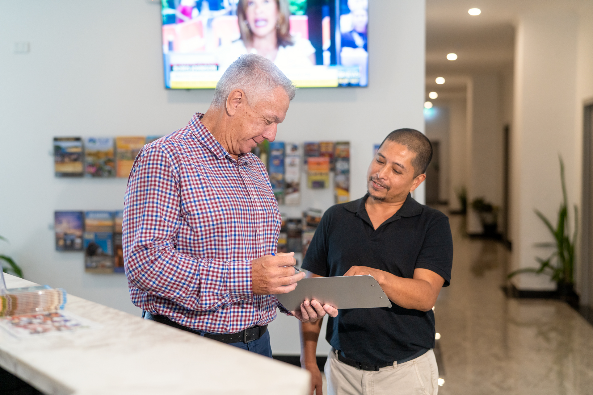 Two people reviewing information on a clipboard at a reception desk in a modern workplace or service centre.