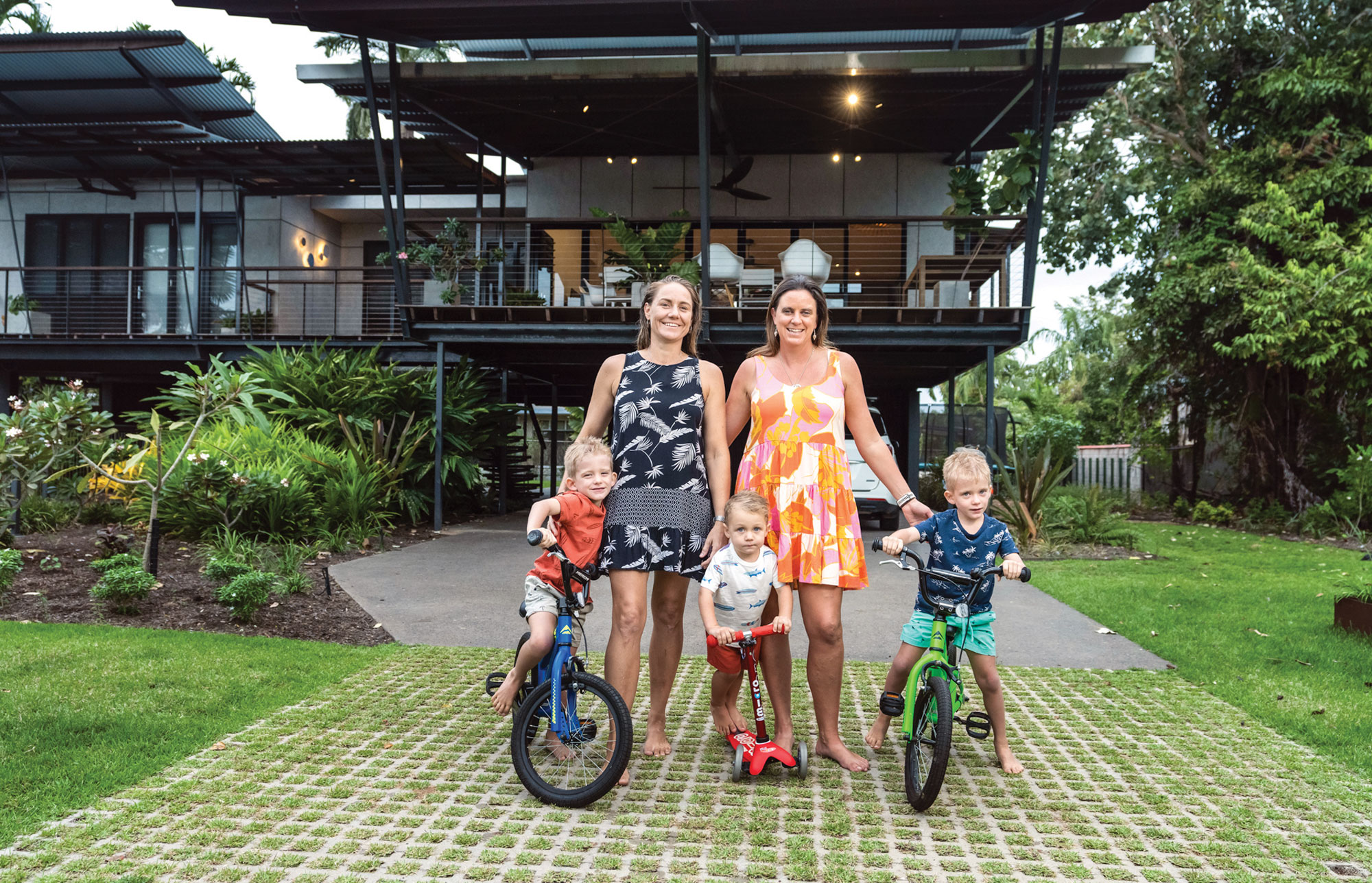 Family with three children on bicycles and a scooter standing in front of a tropical-style home.