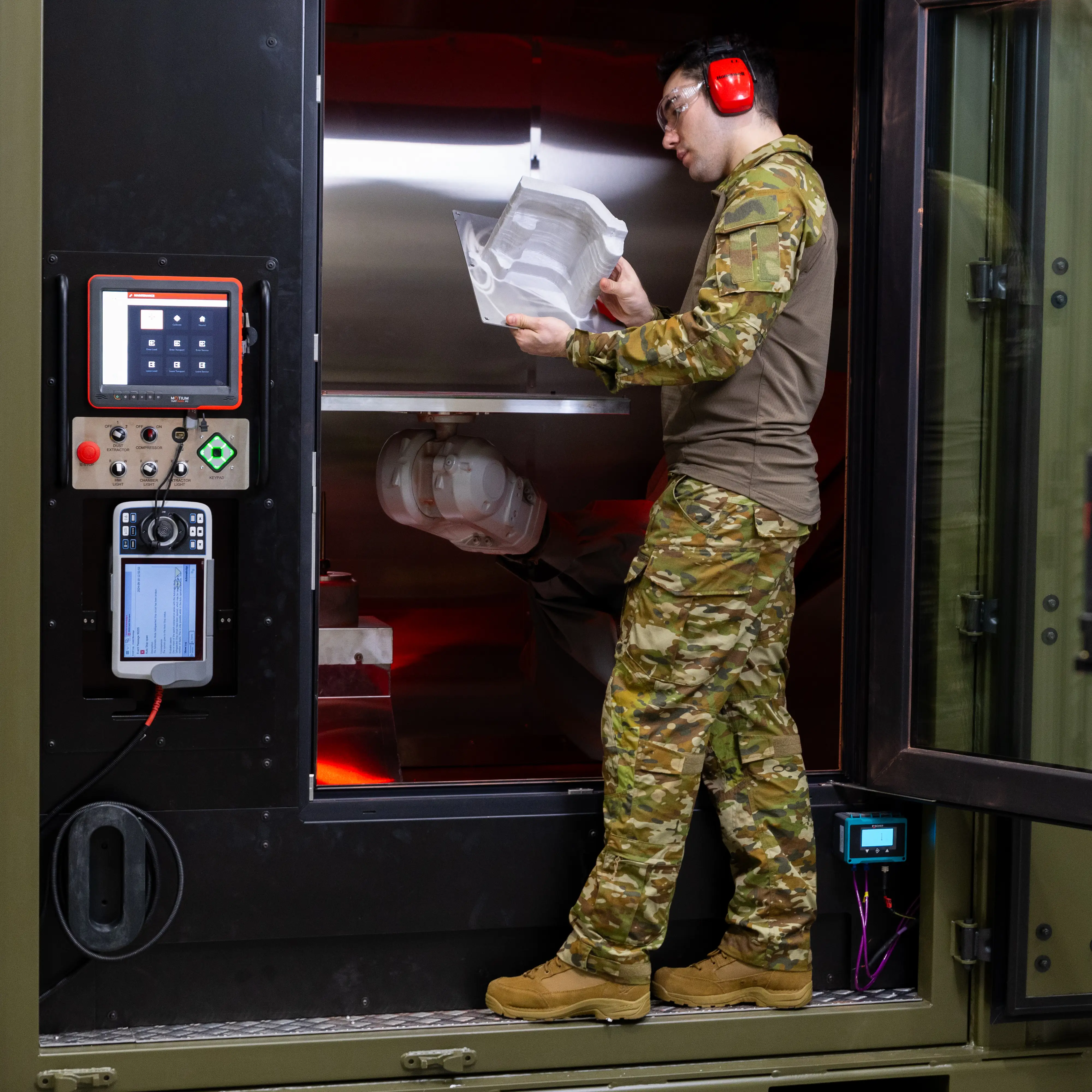 A person wearing camouflage clothing, safety glasses and hearing protection inspects a large manufactured component inside an industrial machine with digital control panels.