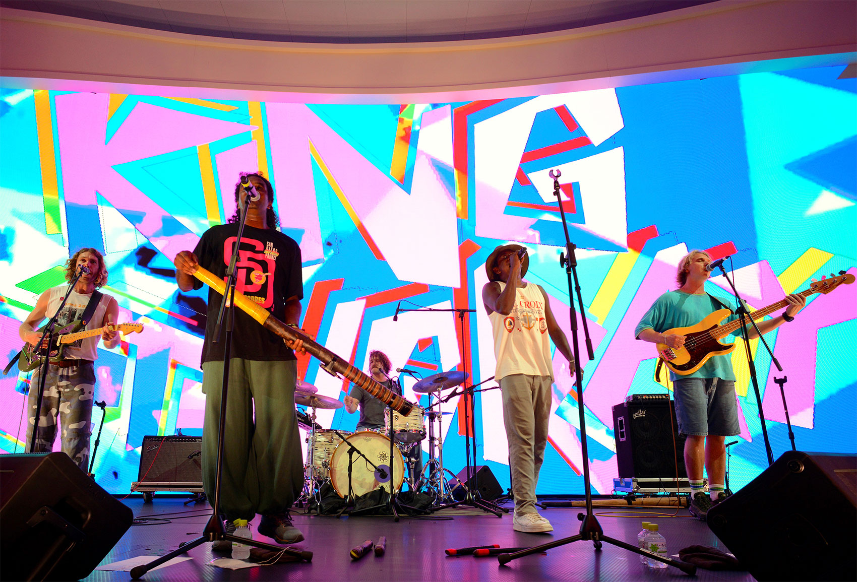 A five-member band performs live on stage with guitars, drums, and vocals, backed by a vibrant LED screen displaying abstract visuals and the word 