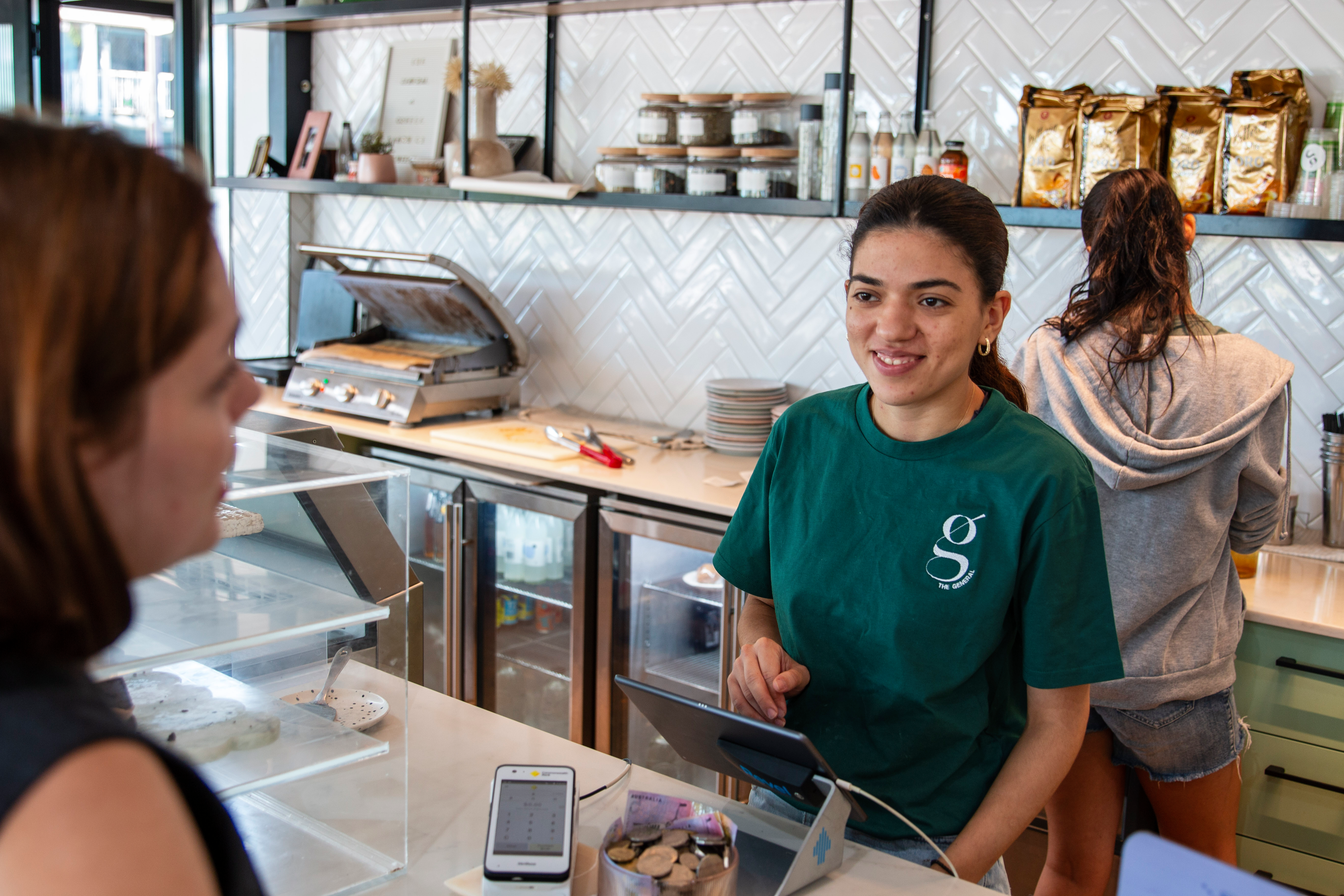 Customer being served at a café counter by a staff member using a point-of-sale terminal while another staff member prepares orders in the background.