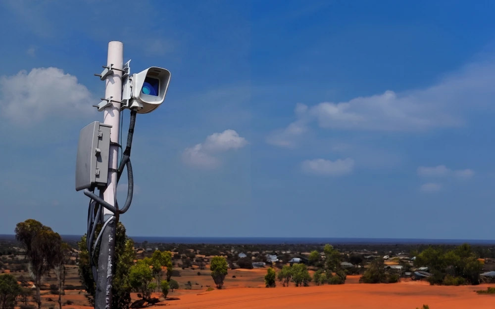 Remote sensing equipment mounted on a pole overlooking a red desert landscape with scattered vegetation and buildings under a blue sky.