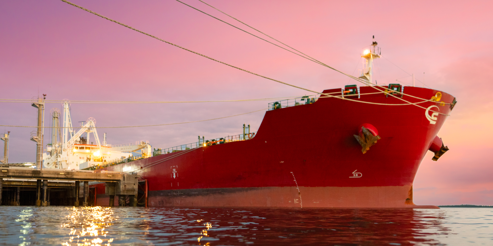 Large red cargo ship docked at a port at sunset, with lights reflecting on the water in the foreground.