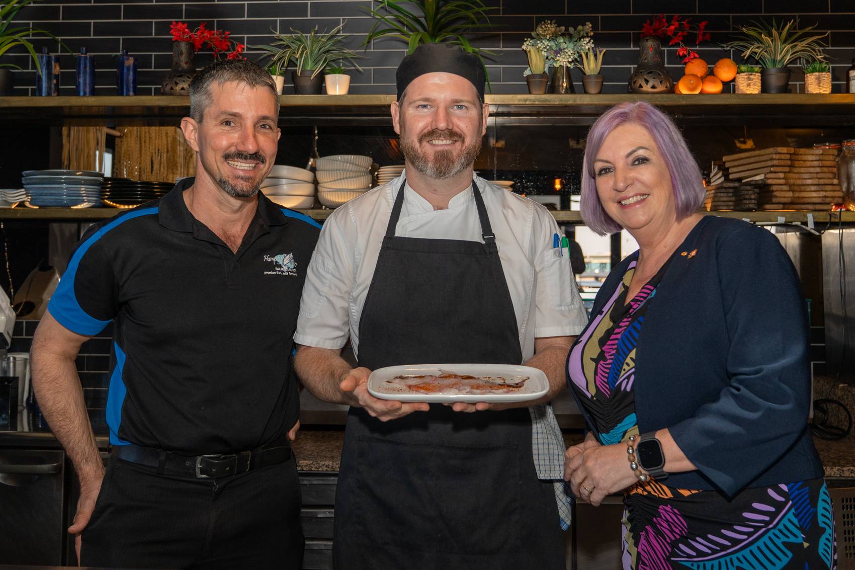Three people smiling in a restaurant kitchen, chef holding a plated dish.