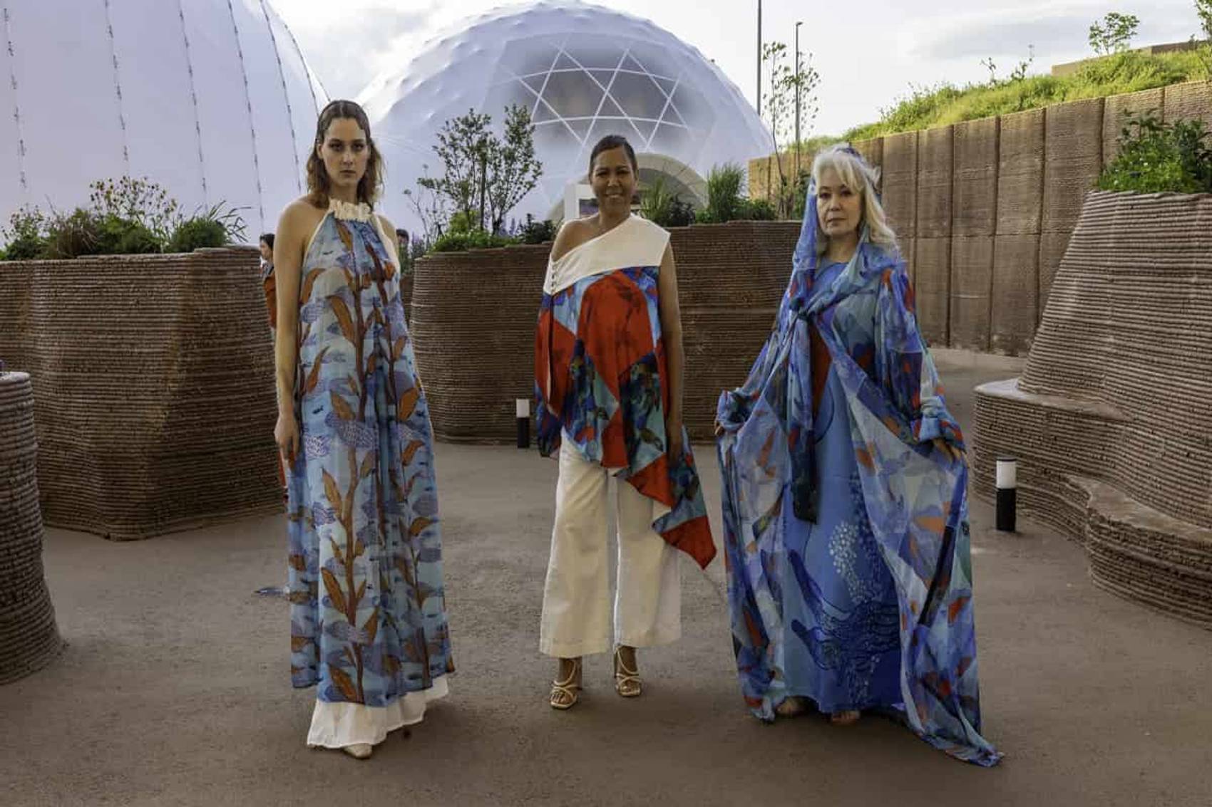 Three people wearing vibrant, flowing garments stand in a futuristic outdoor setting.