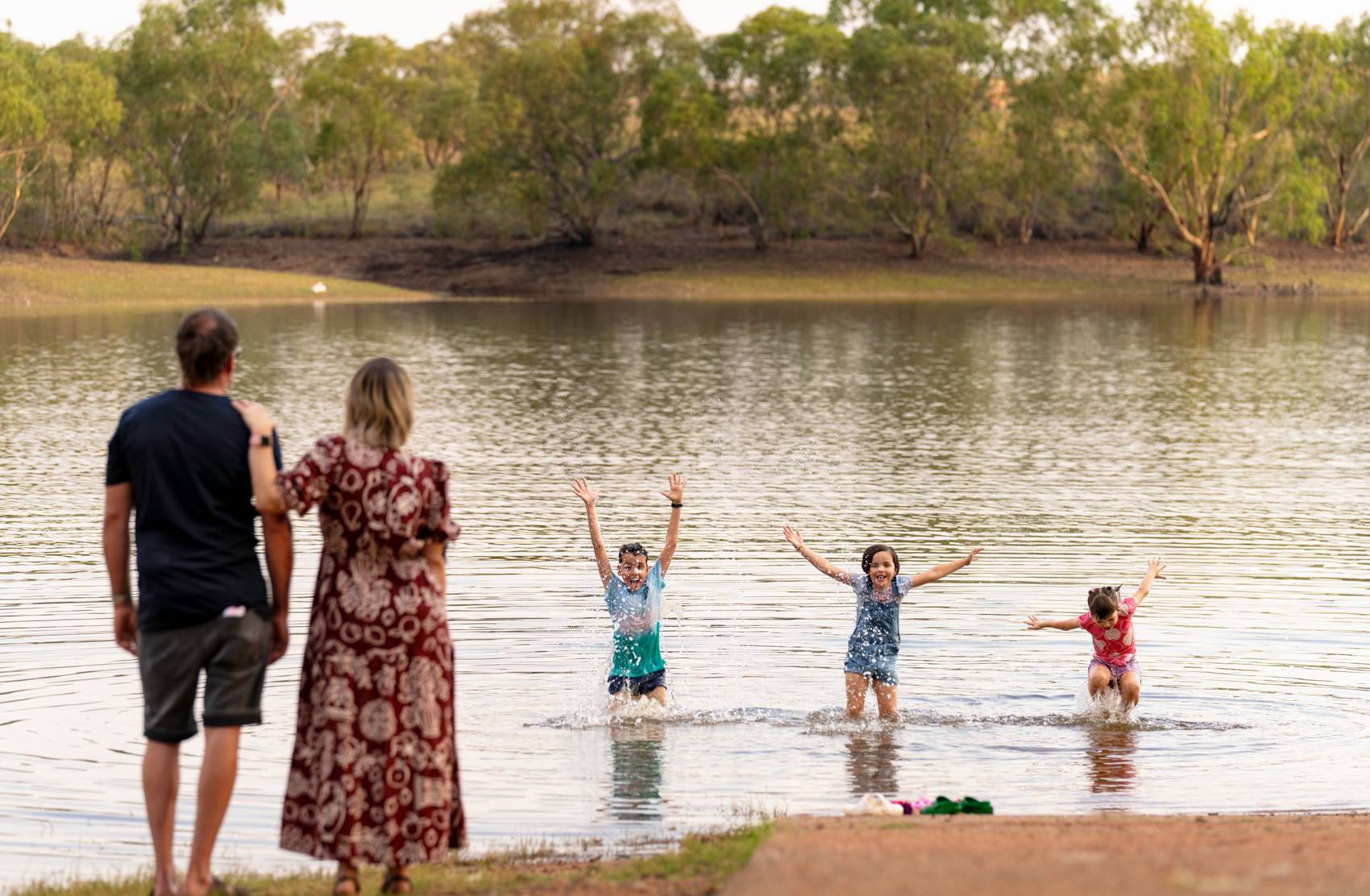 Children joyfully playing in a lake, watched by two adults.