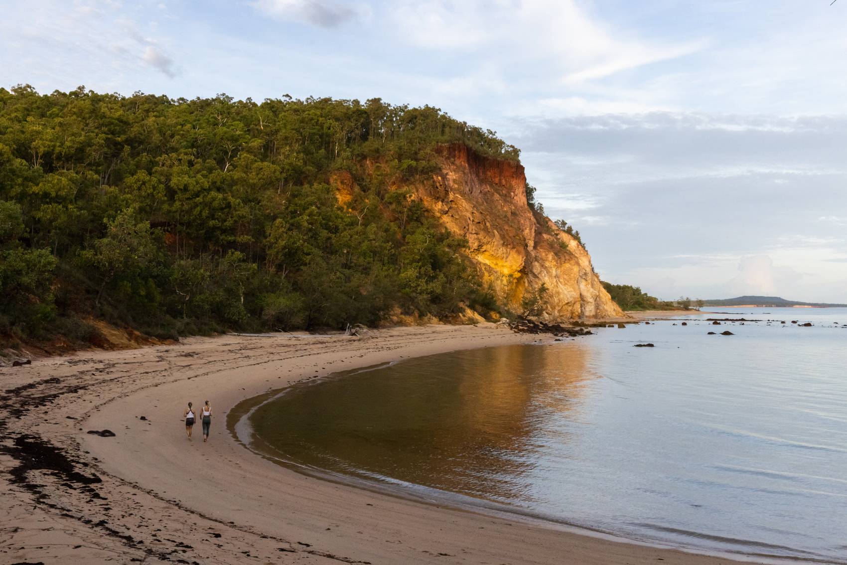 Two people walk along a serene beach with cliffs and lush greenery.