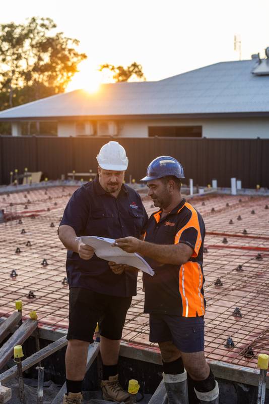 Two construction workers review plans on site at sunset.