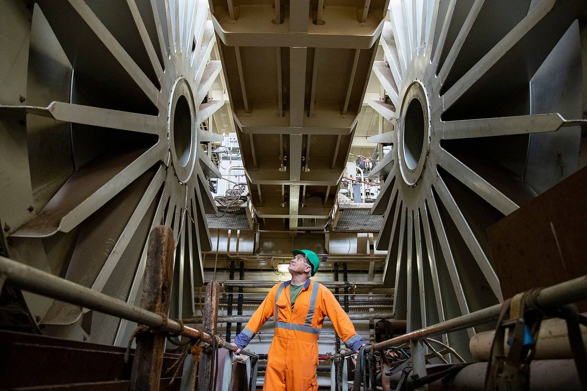 Worker in orange attire inspects large industrial machinery, looking upwards thoughtfully.