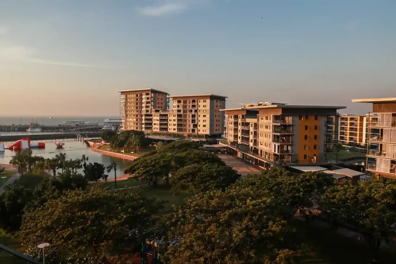 Coastal view of modern apartment buildings at Darwin Waterfront during golden hour, with trees, a swimming lagoon, and inflatable water park in the foreground.